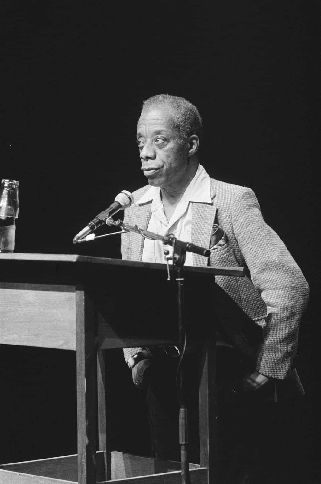James Baldwin speaking at a lectern with a microphone and a bottle beside him, wearing a tweed jacket over an open-necked shirt, Amsterdam 1984