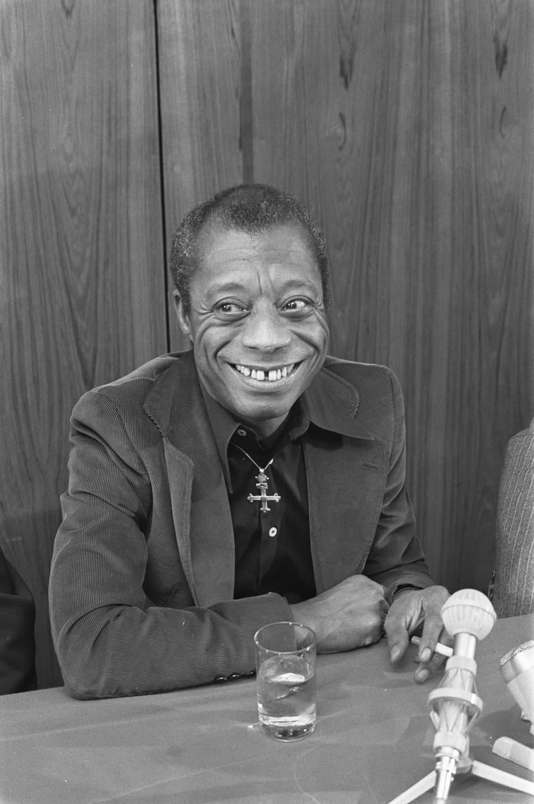 James Baldwin smiling broadly at a press conference table in Amsterdam in 1974, a microphone and a glass of water in front of him, wearing a corduroy jacket over a dark shirt and a metal cross on a chain