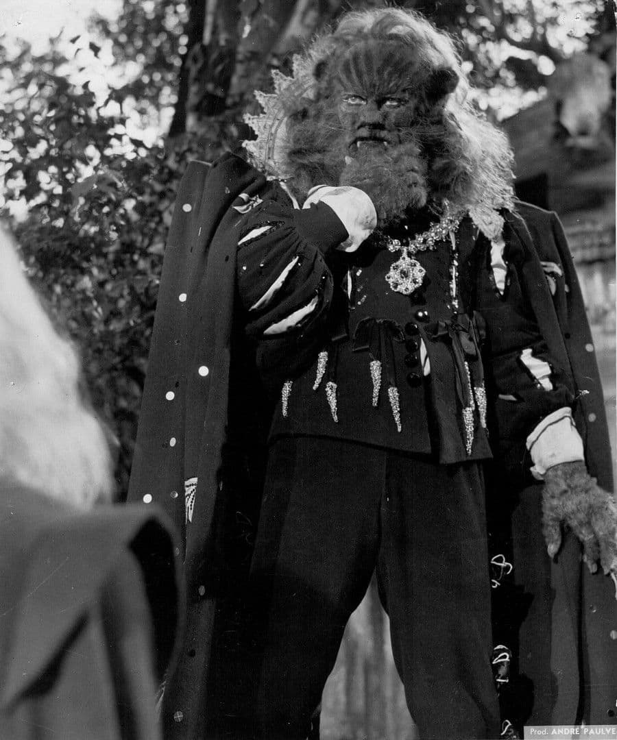 Jean Marais in the full Beast makeup and jeweled court dress, standing outdoors, clawed hand at his side