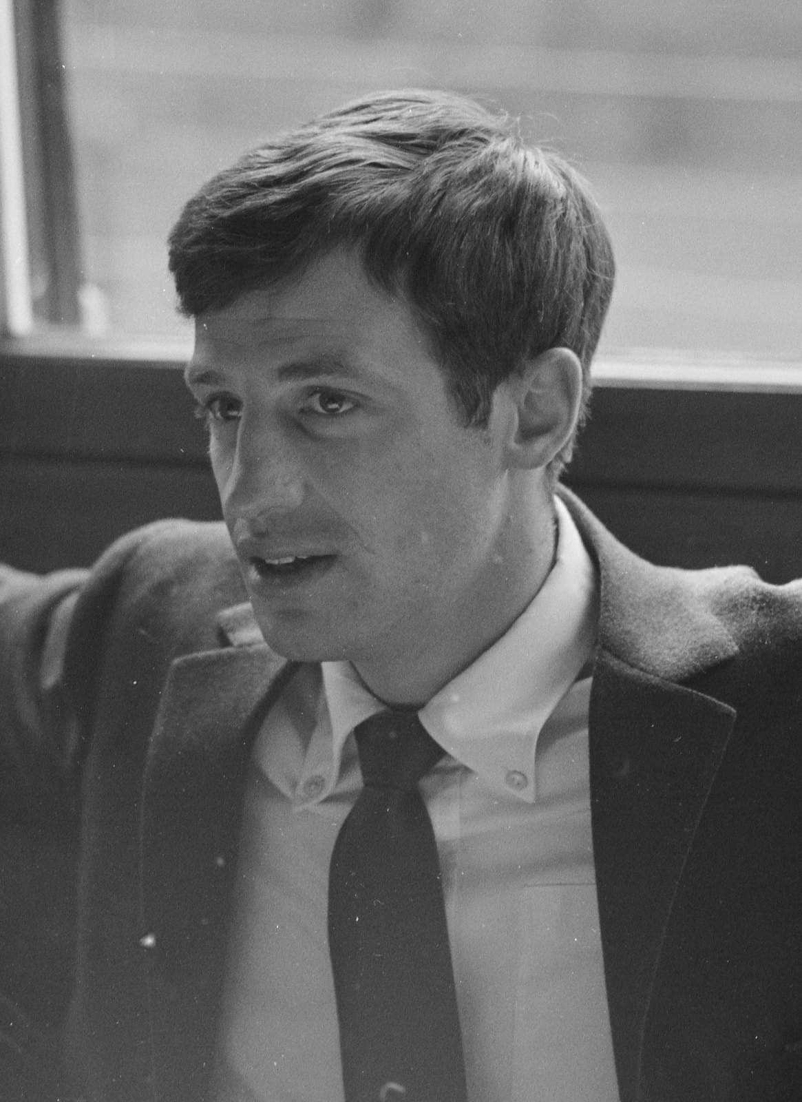 Jean-Paul Belmondo in a jacket and knitted tie, seated by a window, mid-conversation.