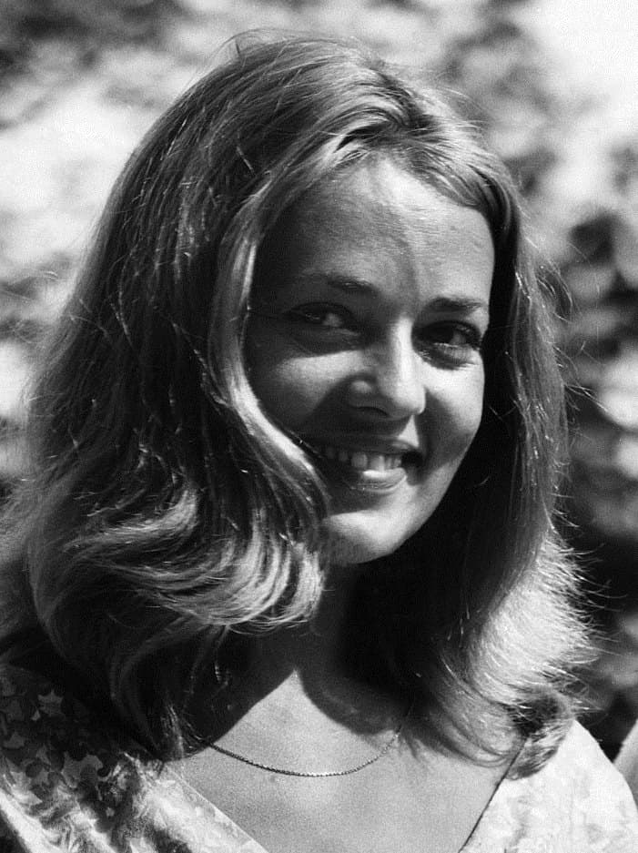 Jeanne Moreau smiling outdoors in dappled light, hair to her shoulders, in a floral dress.