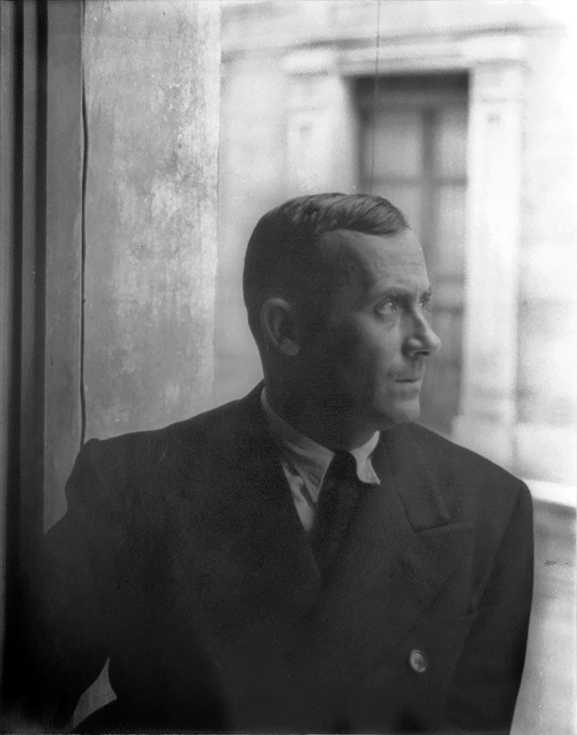 Joan Miró in a dark suit beside a window, looking to the right against a courtyard wall.