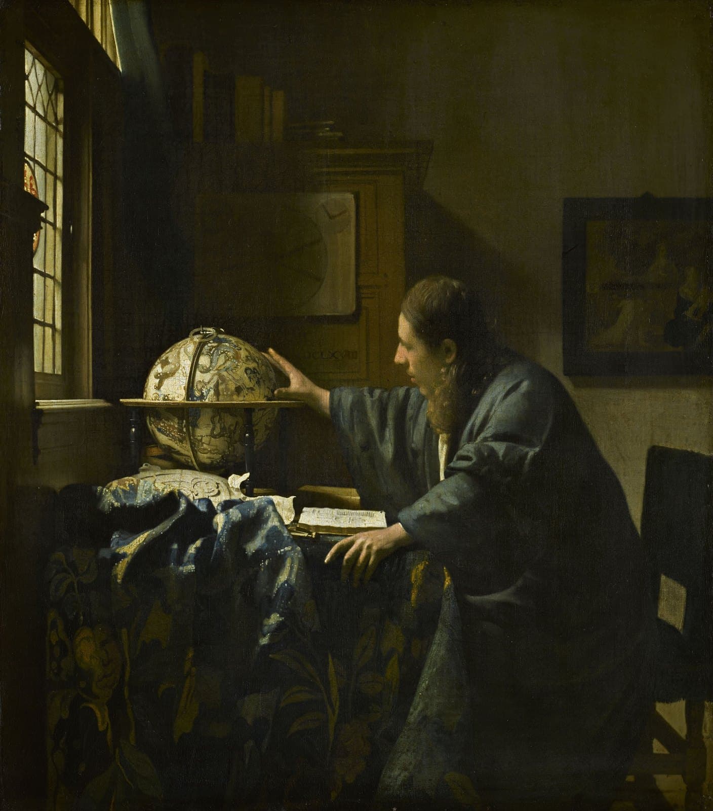 A man with long hair in a loose green-blue gown sits at a table covered with a patterned cloth and reaches toward a celestial globe. An open book lies in front of him, light comes from a window at the left, and a framed painting hangs on the wall behind him.