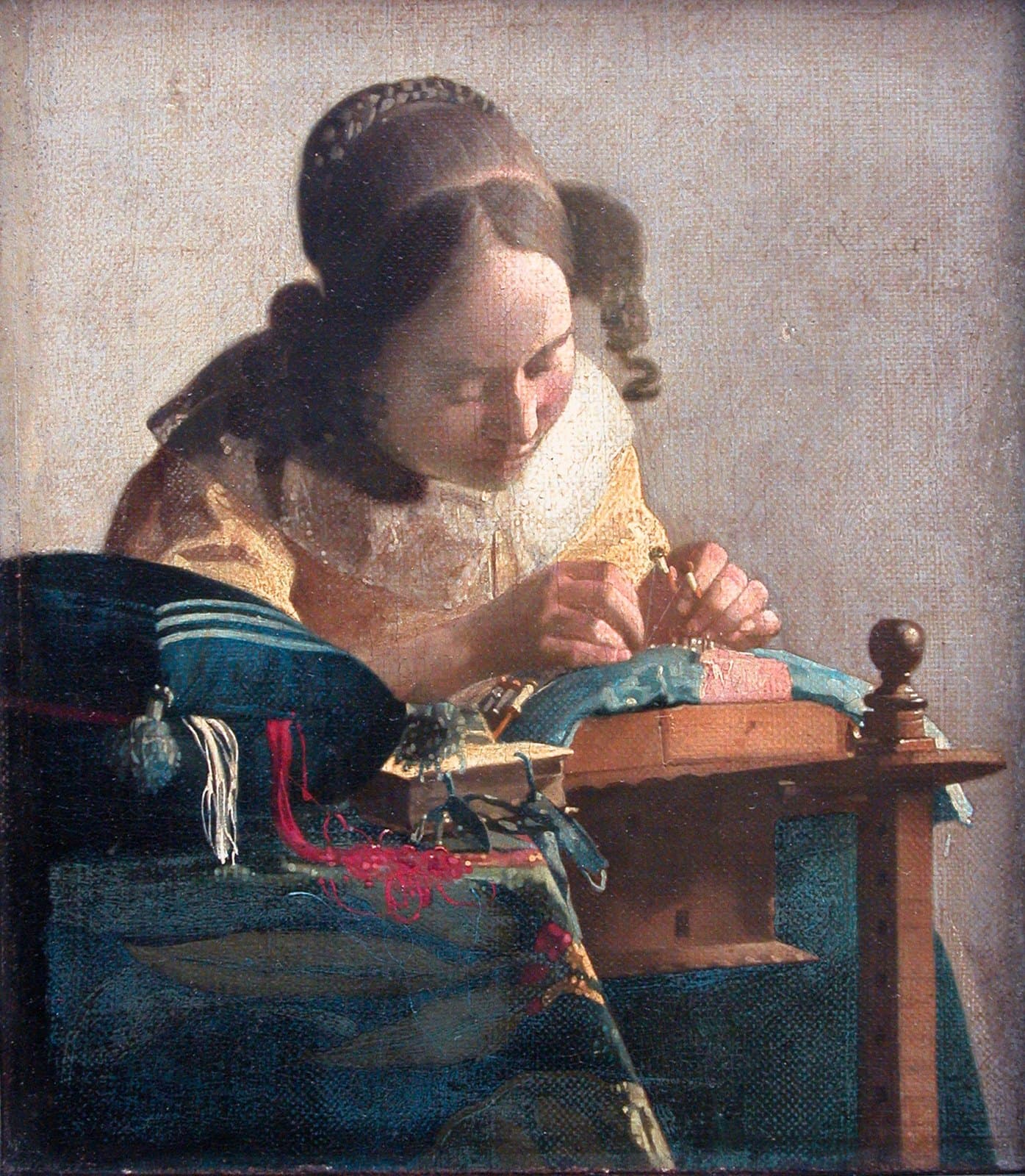 A young woman in a yellow bodice bends over a lace pillow, holding bobbins in both hands. A blue sewing cushion with red and white threads spilling from it sits on the table in front of her, next to a small book.