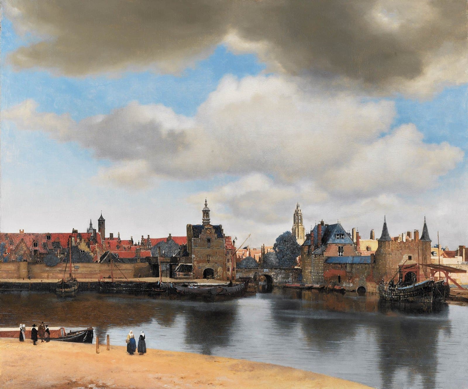 A town seen across calm water under a wide sky of gray and white clouds. Brick gates, red roofs, and a church tower lit by sunlight line the far bank, boats are moored at the quay, and a few small figures stand on the sandy near shore.