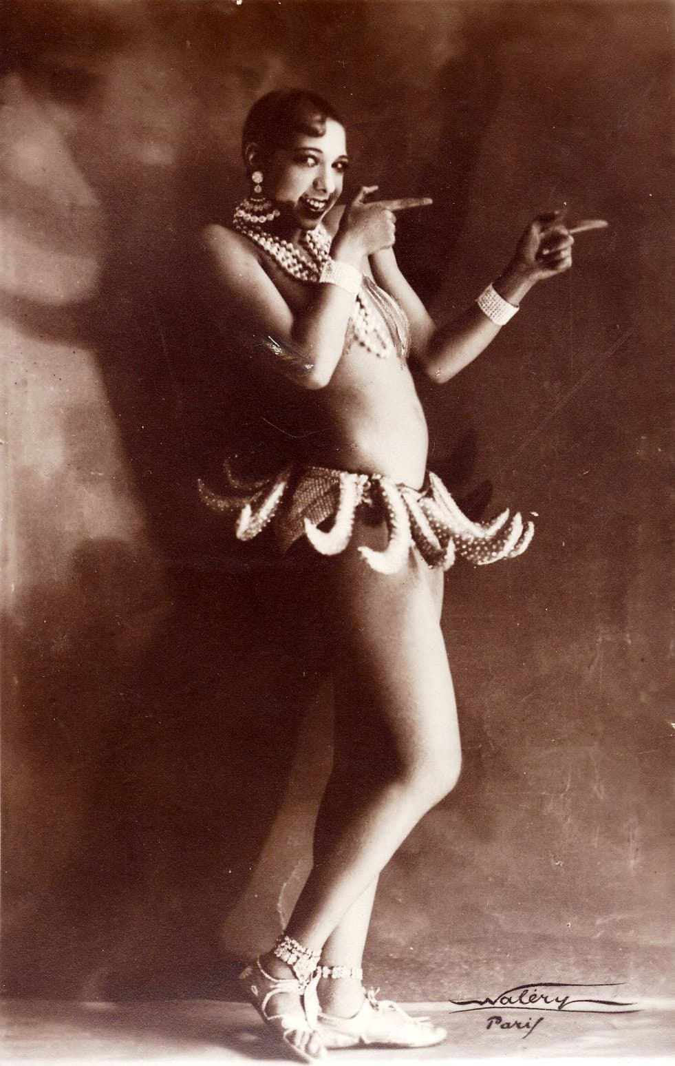 Josephine Baker in the banana skirt, hands on her hips, beaded necklace, photographed against a dark studio background