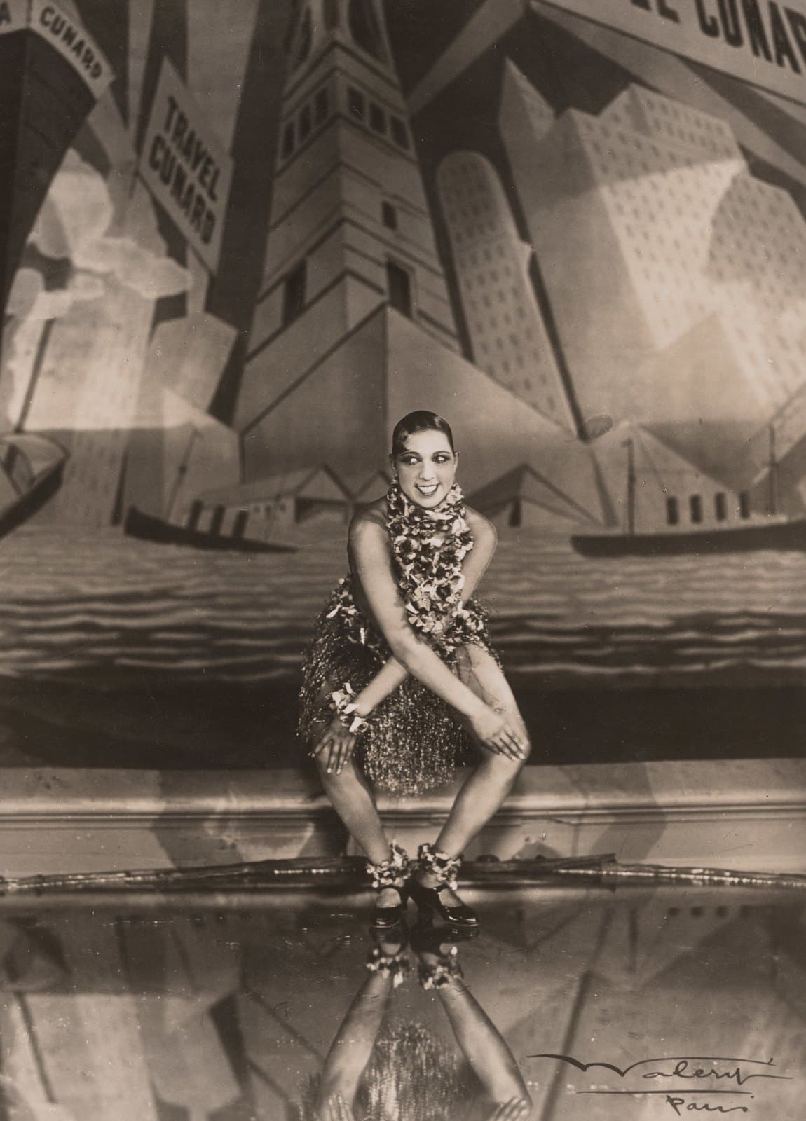 Josephine Baker mid-Charleston in a fringed skirt, knees bent inward, arms crossed, grinning at the camera
