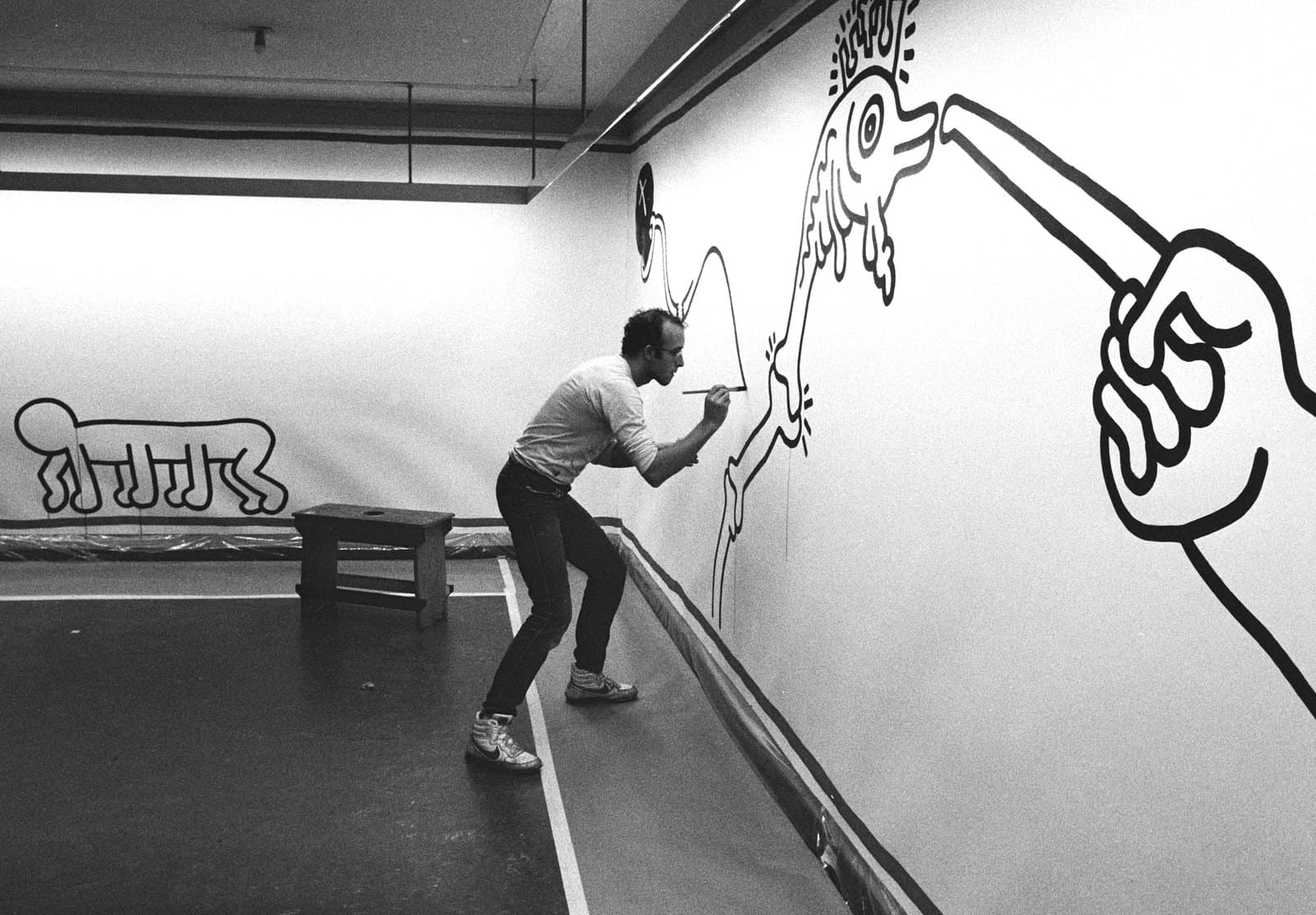 Keith Haring painting a large black outlined figure on a white wall at the Stedelijk Museum in 1986.