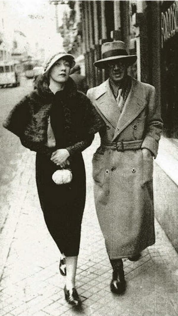 Kiki de Montparnasse in a cloche hat and fur-trimmed coat walking a Paris street beside Tsuguharu Foujita, who wears a belted overcoat and round glasses.