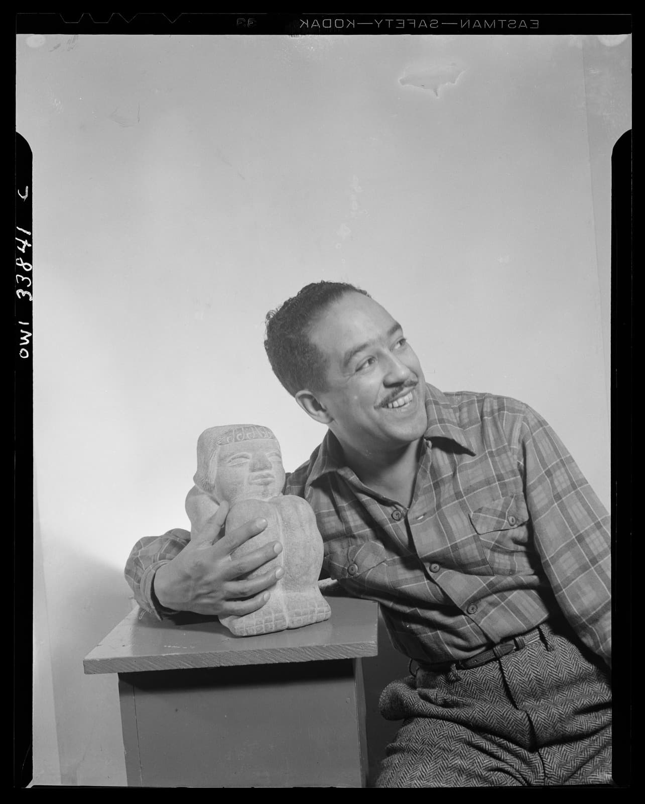 Portrait of Langston Hughes in 1943, seated beside a sculpture and smiling toward the camera