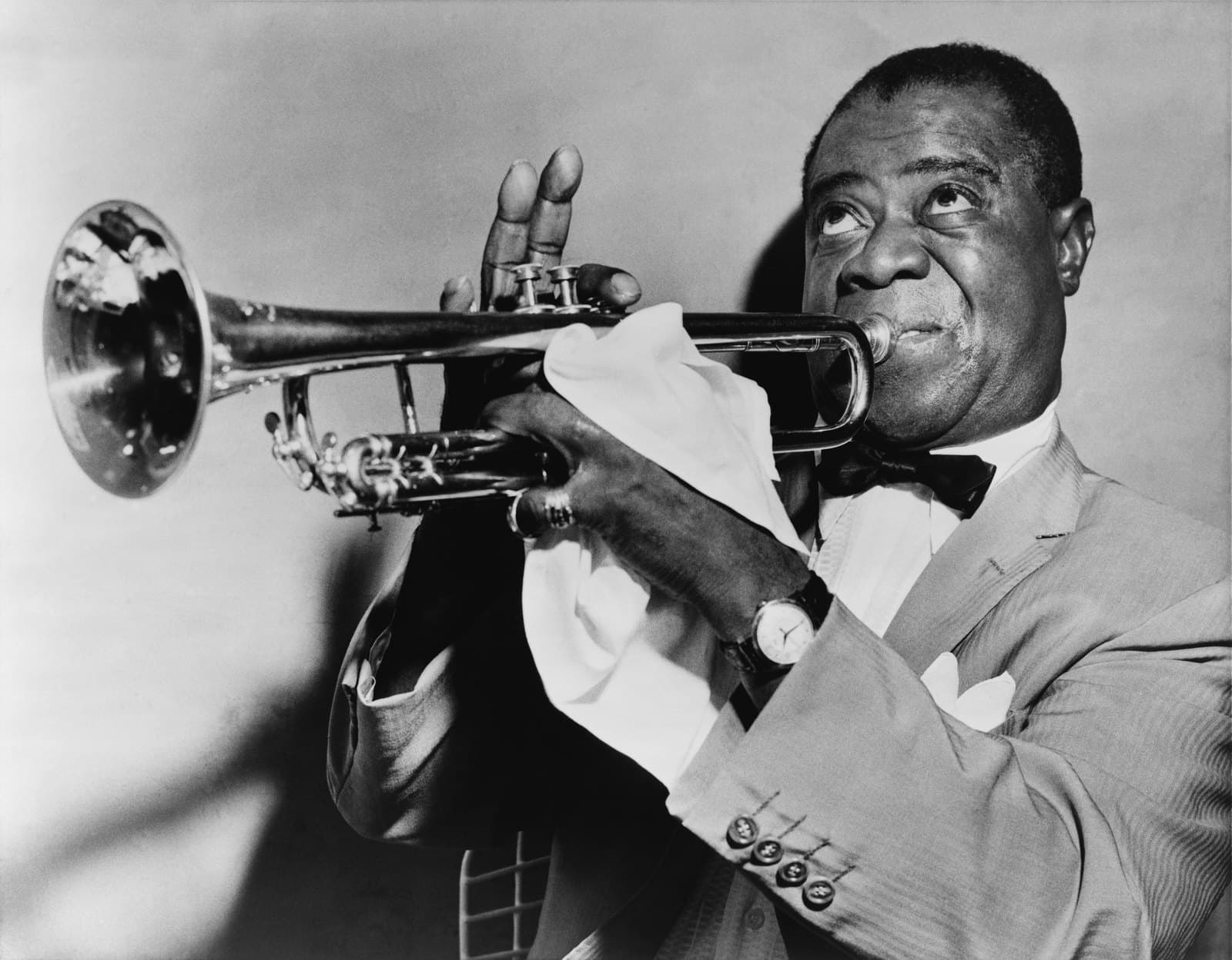 Louis Armstrong in 1953, playing trumpet in a dark suit with a handkerchief tucked beneath the mouthpiece.