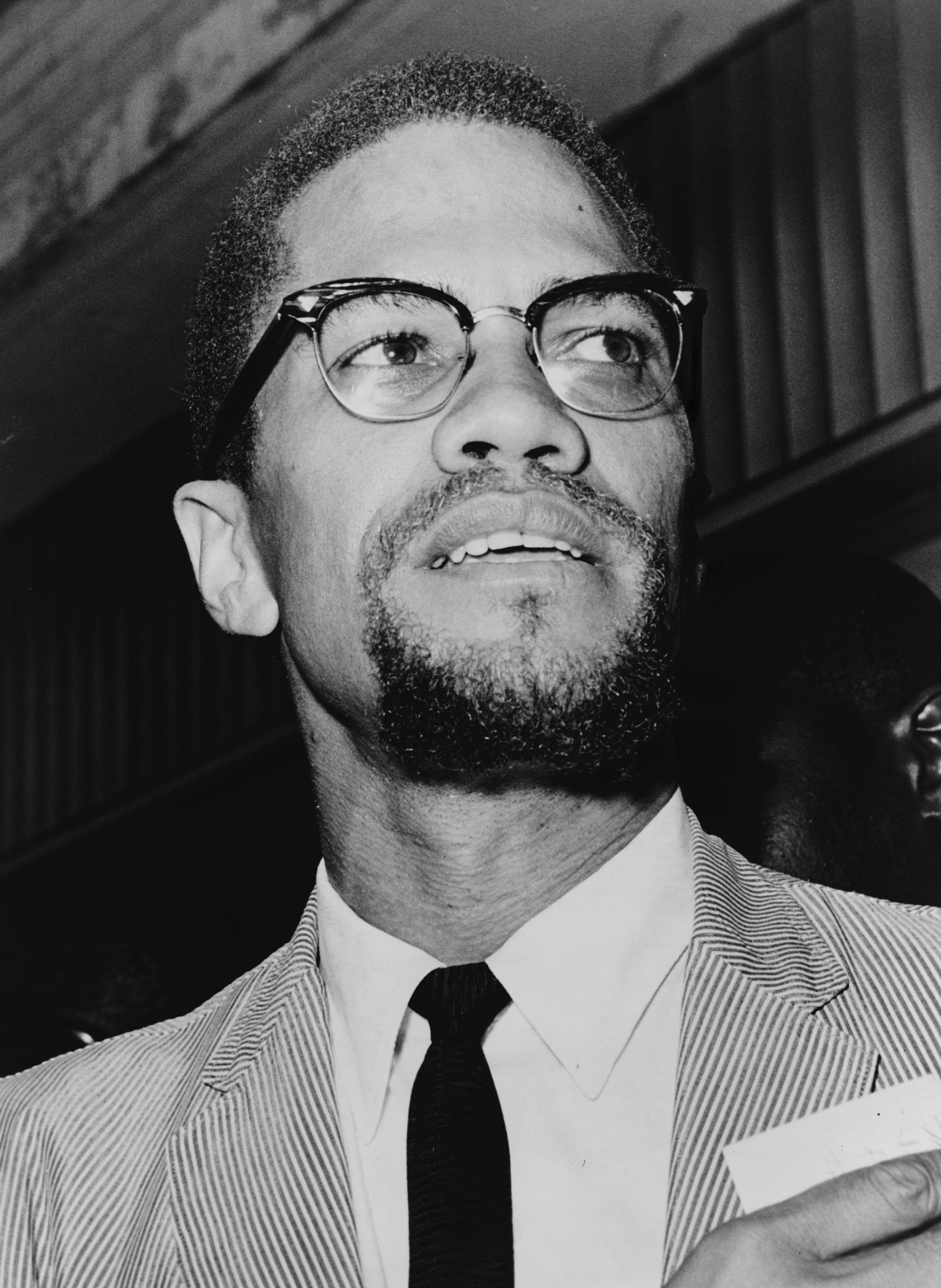 Malcolm X in a dark suit and horn-rimmed glasses, seated and looking off to one side.
