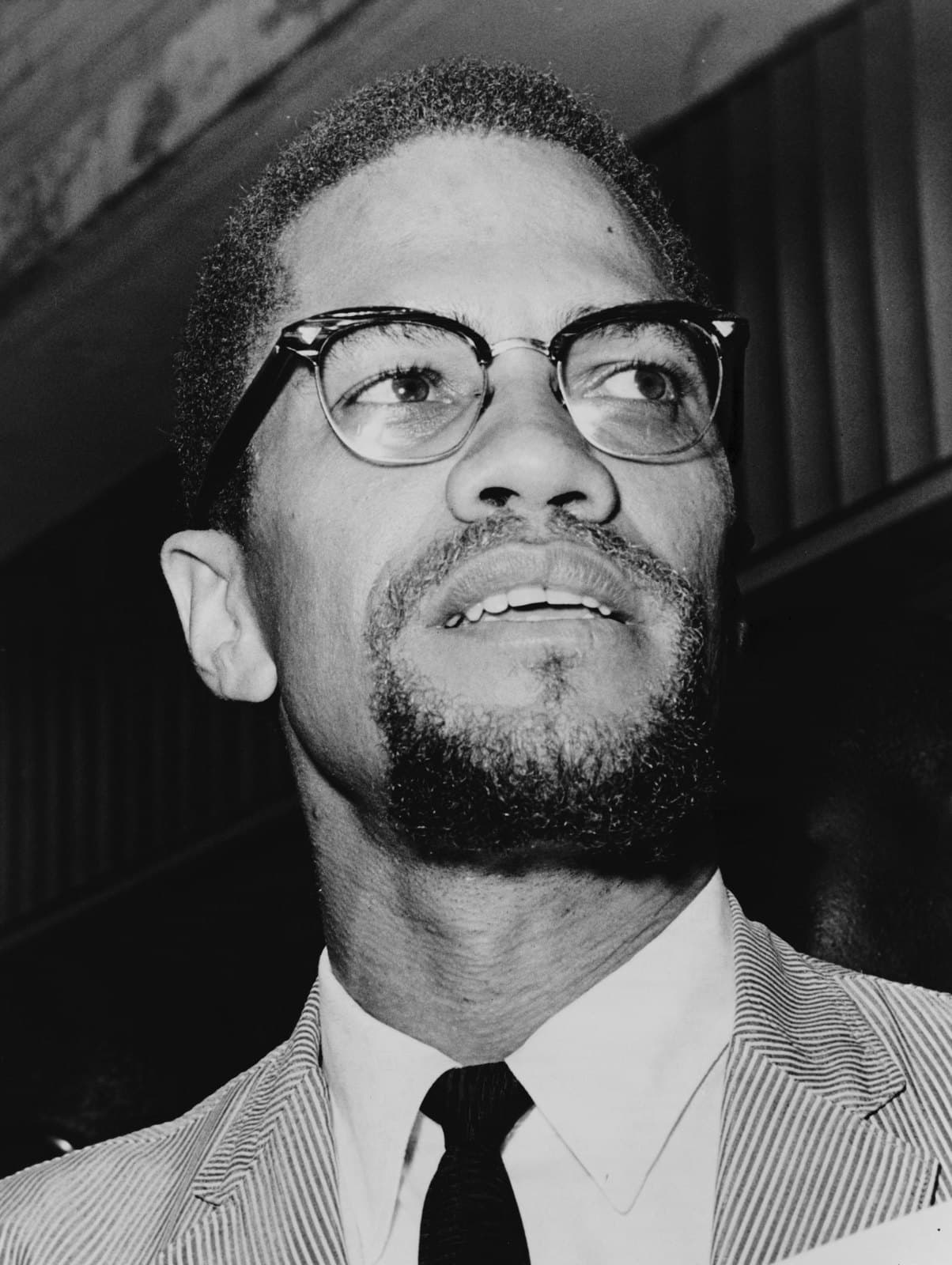 Malcolm X in profile wearing dark-rimmed glasses and speaking to reporters in 1964.