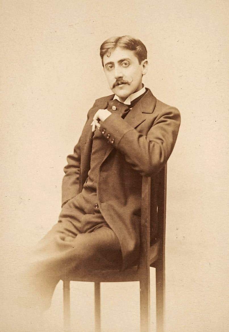 Marcel Proust in his mid-twenties seated on a chair, one arm over its back, in a dark suit.