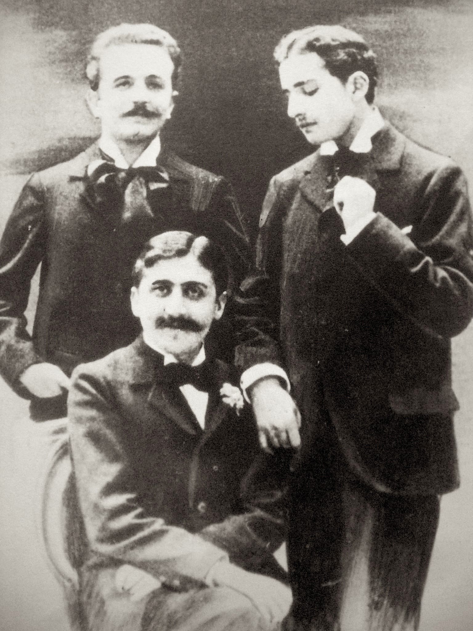 Marcel Proust seated between two standing friends: Robert de Flers at left, Lucien Daudet at right leaning on his chair.