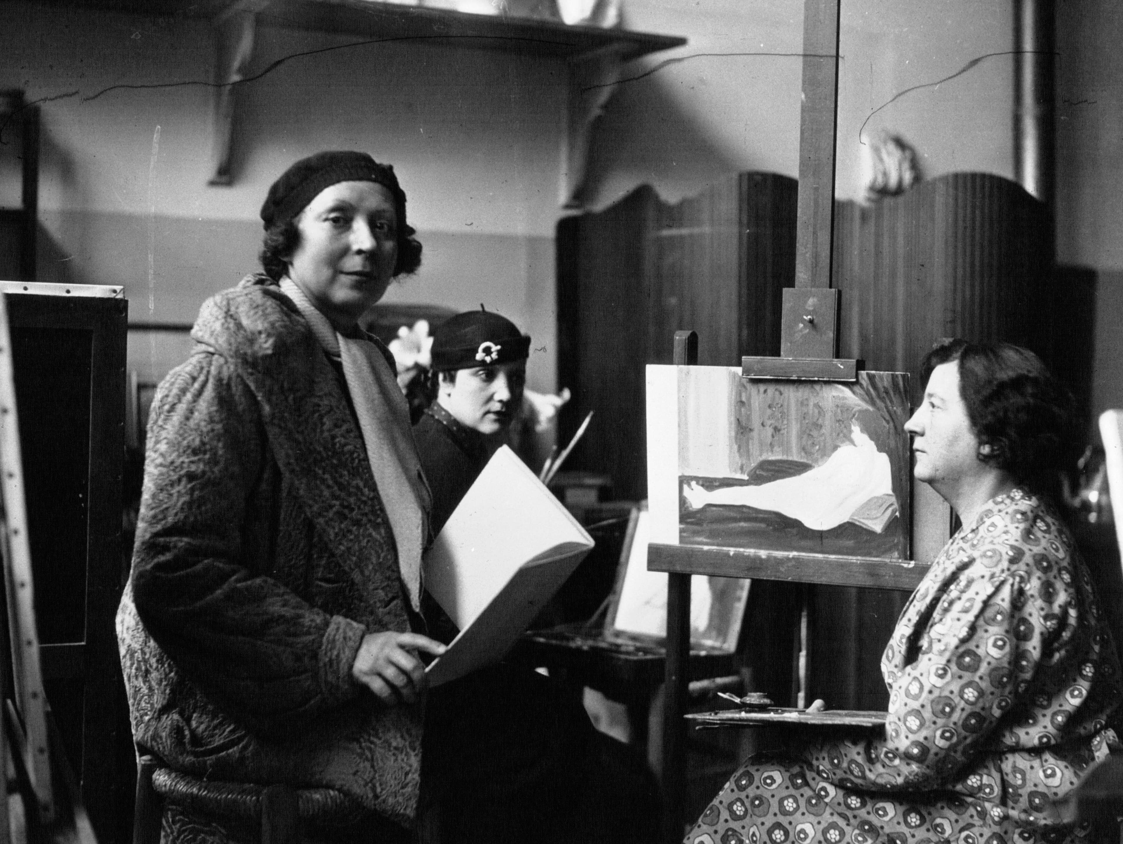 Marie Laurencin standing in her atelier in 1932, holding a portfolio, with a student behind her and a sitter posed beside a painting on an easel