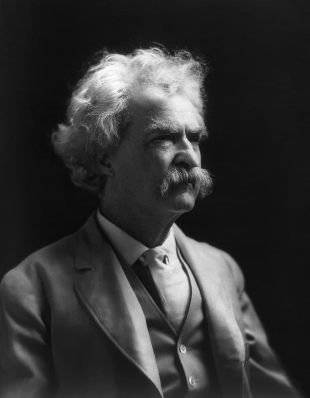 Mark Twain in a 1907 portrait, seated in a white suit with one hand resting against his cheek.