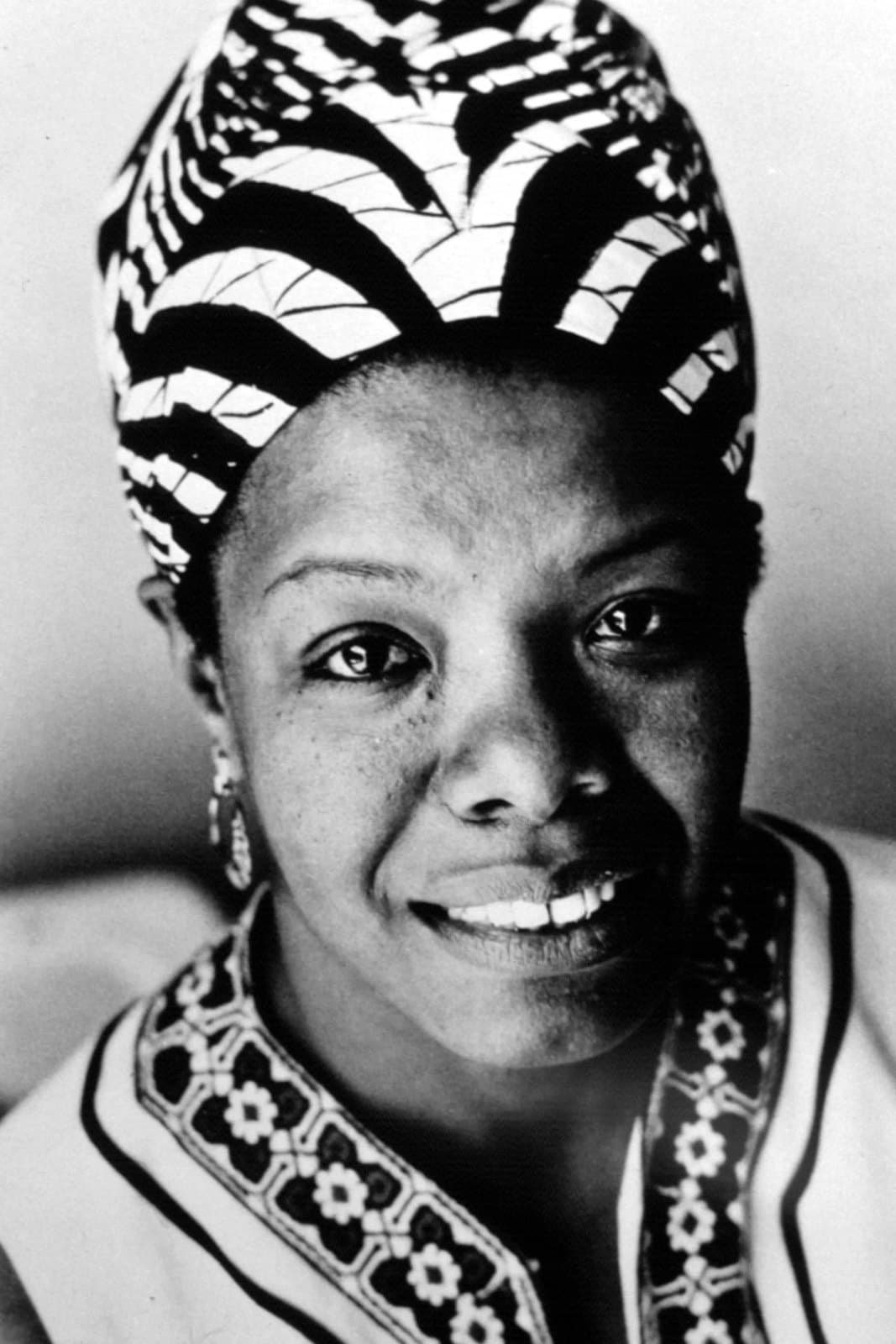 Black-and-white close-up portrait of Maya Angelou smiling, wearing a boldly patterned head-wrap and an embroidered collar, with drop earrings