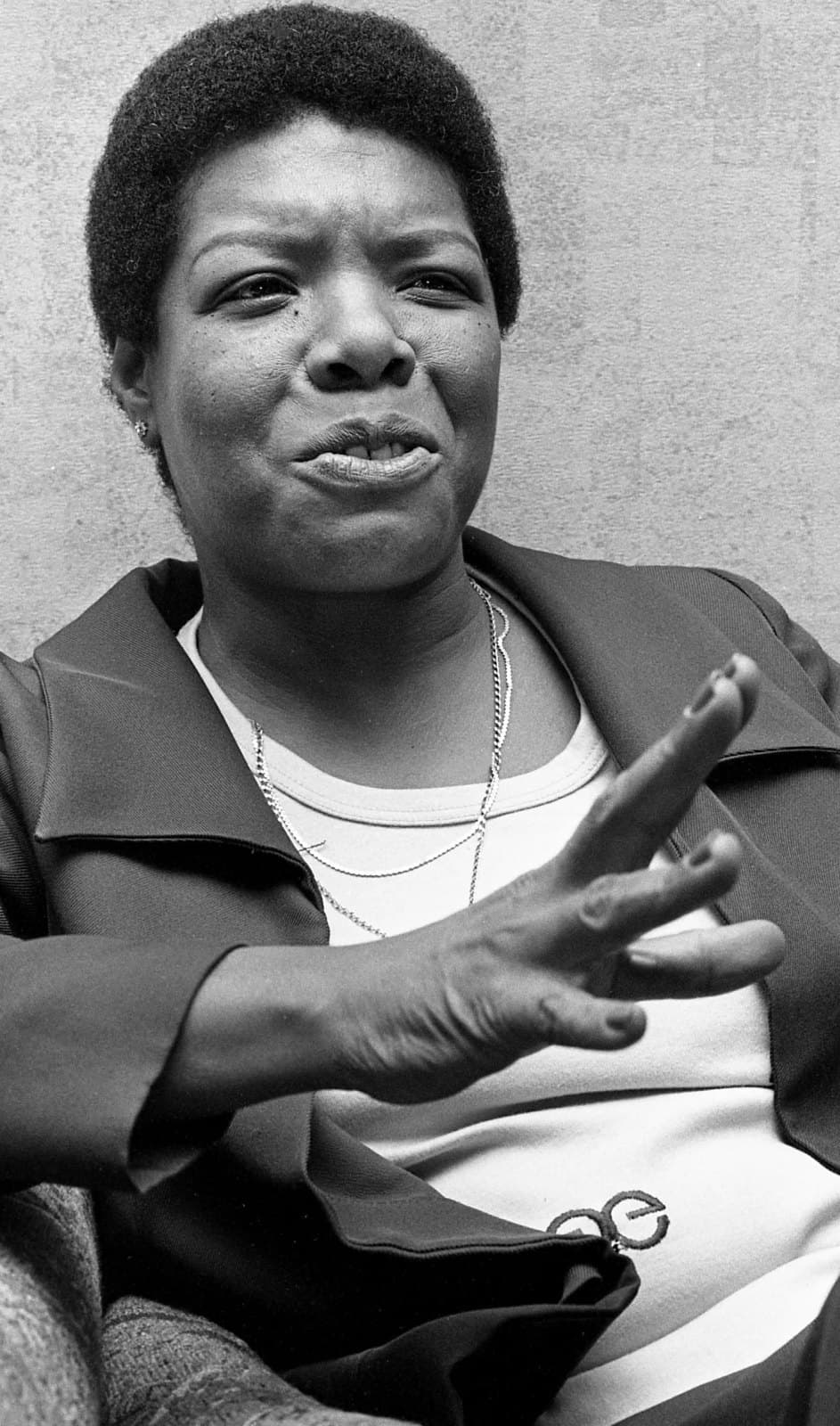 Black-and-white photograph of Maya Angelou mid-conversation, one hand raised in a gesture, speaking, wearing a dark jacket over a white top with necklaces