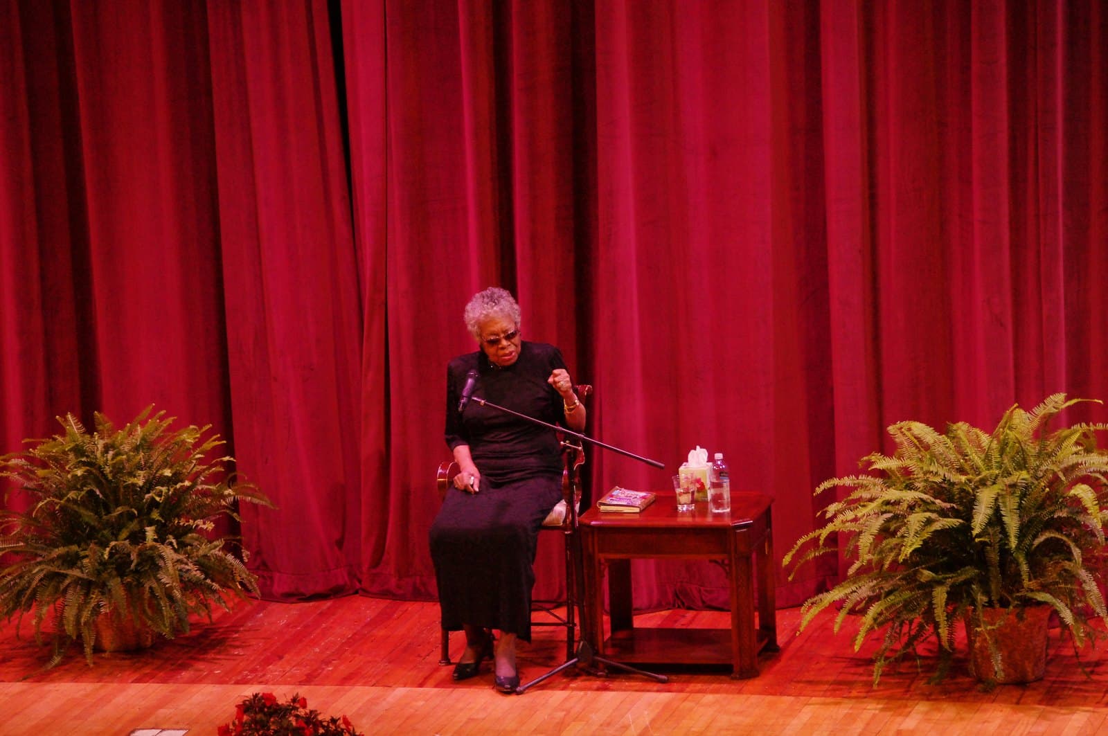 Maya Angelou seated on a stool on a stage before a deep red curtain, speaking into a microphone, with potted ferns and a small table beside her