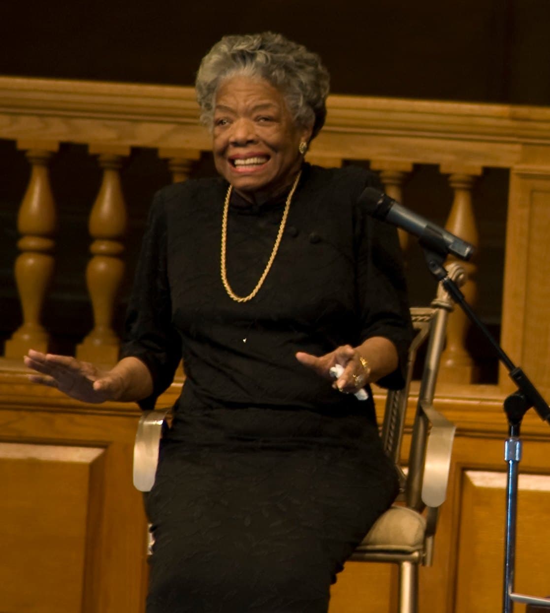 Maya Angelou seated at a microphone, smiling broadly with both hands open in a gesture, before a wooden balustrade