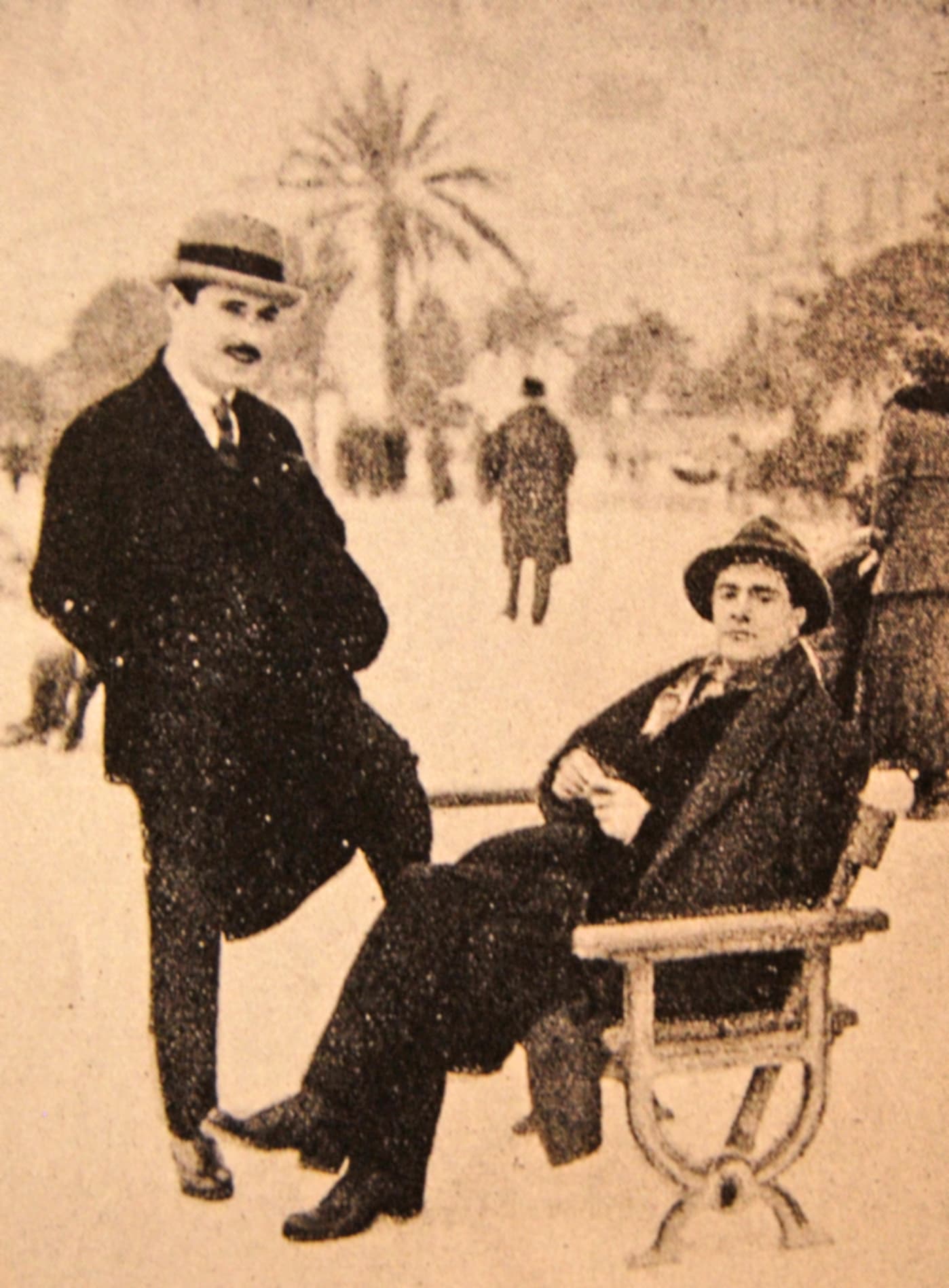 Paul Guillaume standing in a suit and boater beside Amedeo Modigliani, seated on a bench on the Promenade des Anglais in Nice, a palm tree behind them