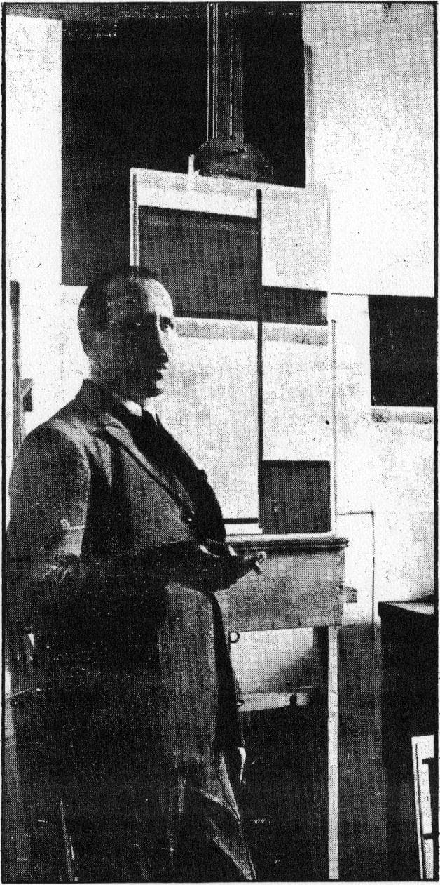 Mondrian standing in his Paris studio in a suit, surrounded by walls hung with rectangular panels and framed grid compositions