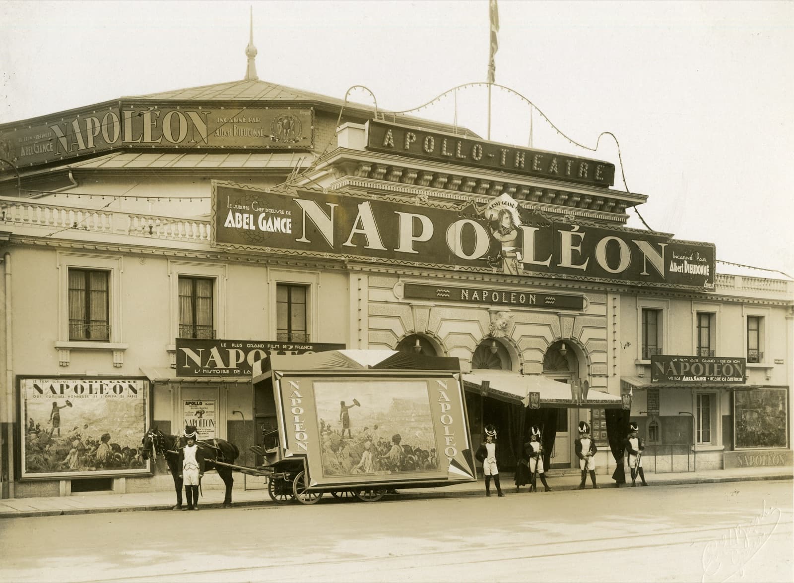 The Apollo-Théâtre facade covered in Napoléon signage, with costumed Napoleonic soldiers and a horse-drawn poster wagon outside