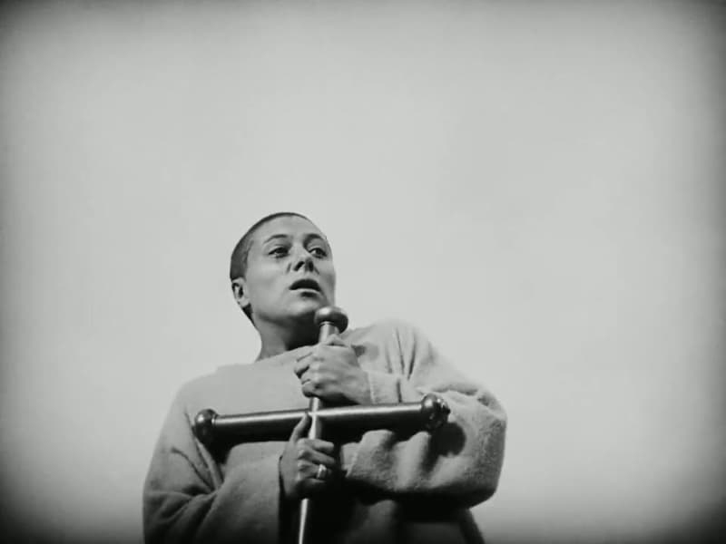Joan, head shaved, clutches a small improvised cross and looks upward