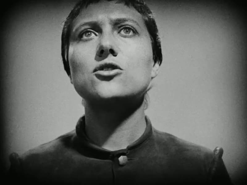 Joan in a dark soldier's tunic looks upward, hair still uncut, during the opening interrogation