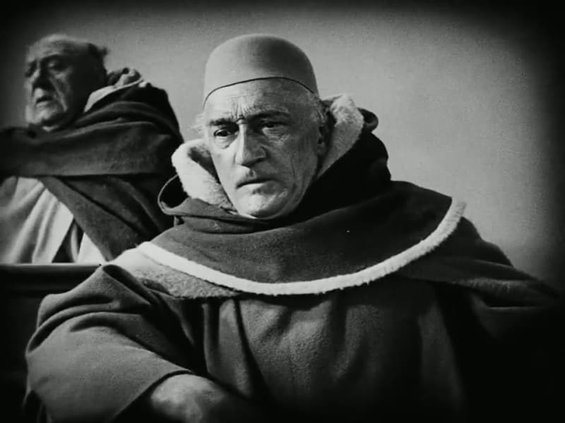 Two judges of the tribunal lean together in close-up, one in an ermine-trimmed robe