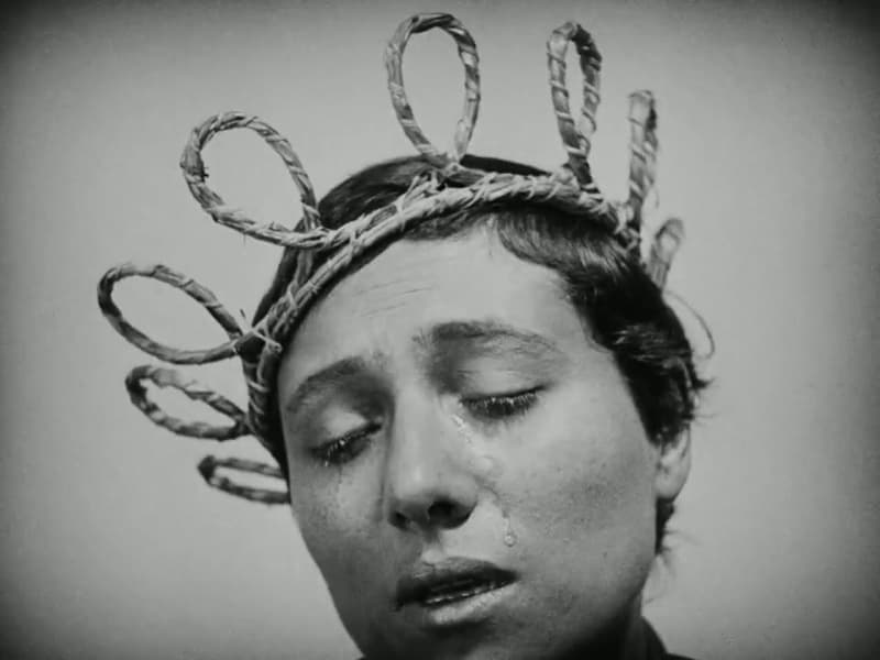 Joan wears a mock crown of plaited straw, eyes closed, tears on her face