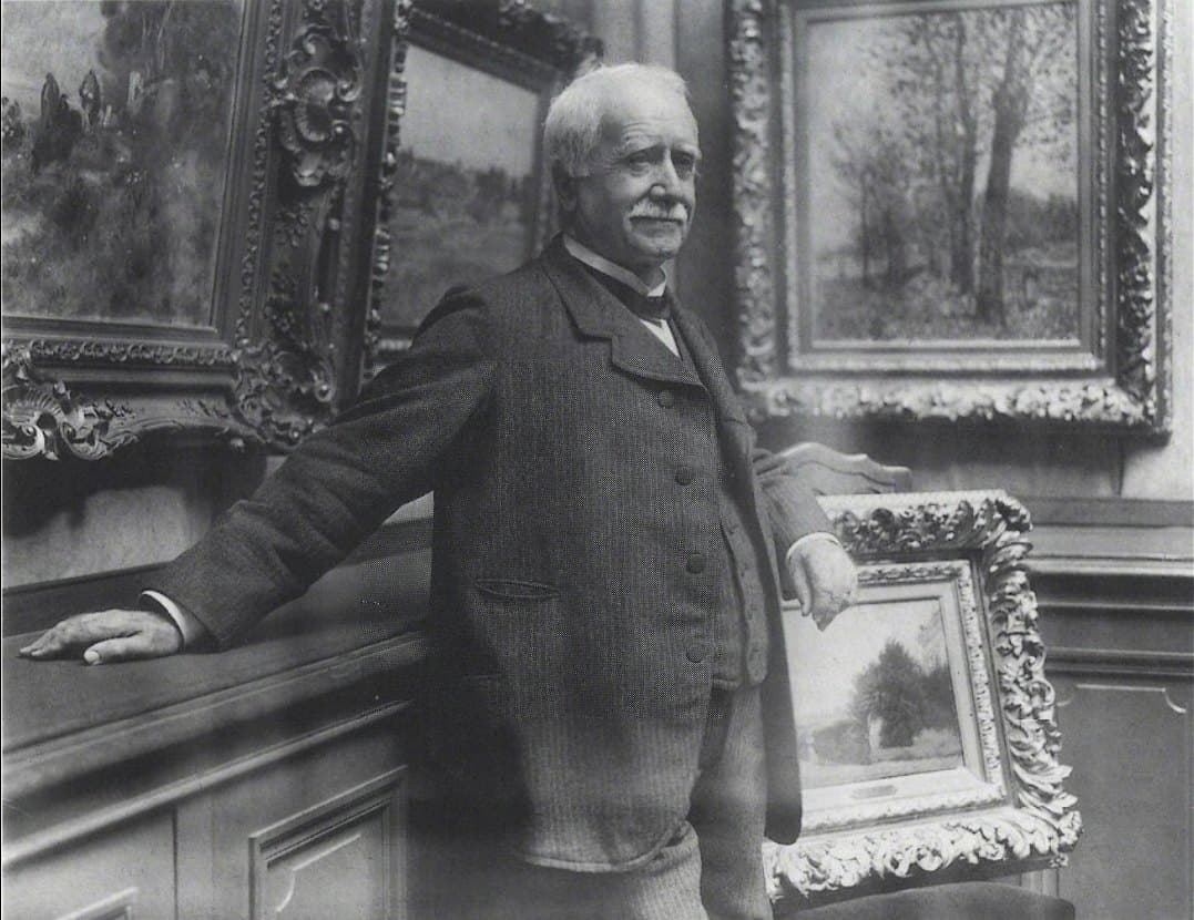 Paul Durand-Ruel, white-moustached in a buttoned coat, leaning against a rail in his picture-hung gallery.