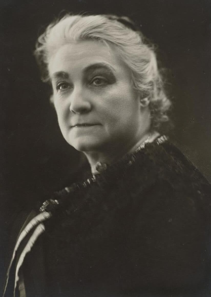 Rachilde in her sixties, head and shoulders against a dark ground, white hair swept back from her forehead, wearing a dark patterned jacket with a ruffled collar.