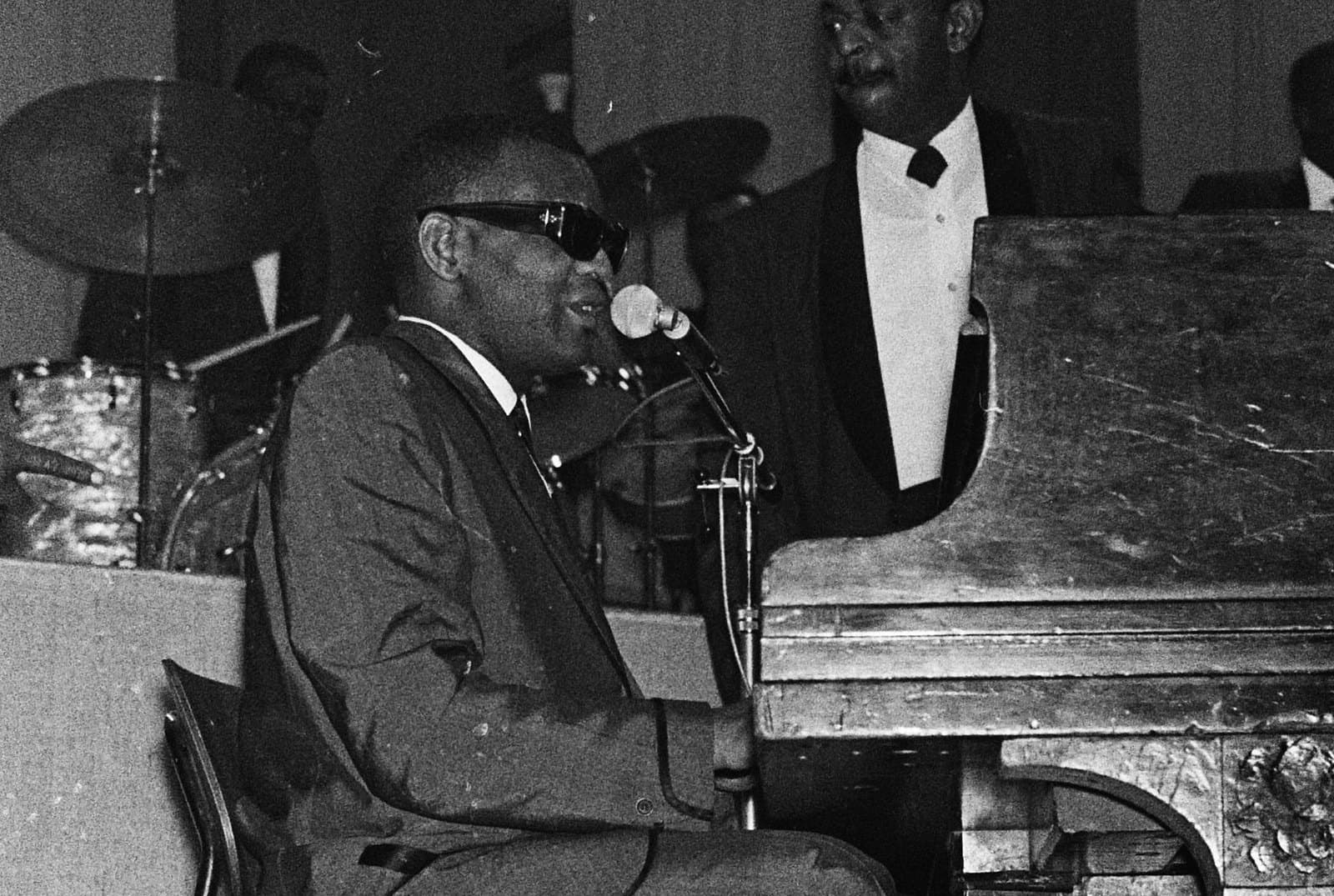 Ray Charles seated at a piano and singing into a microphone during a nighttime concert in Amsterdam in 1963, with members of his band behind him