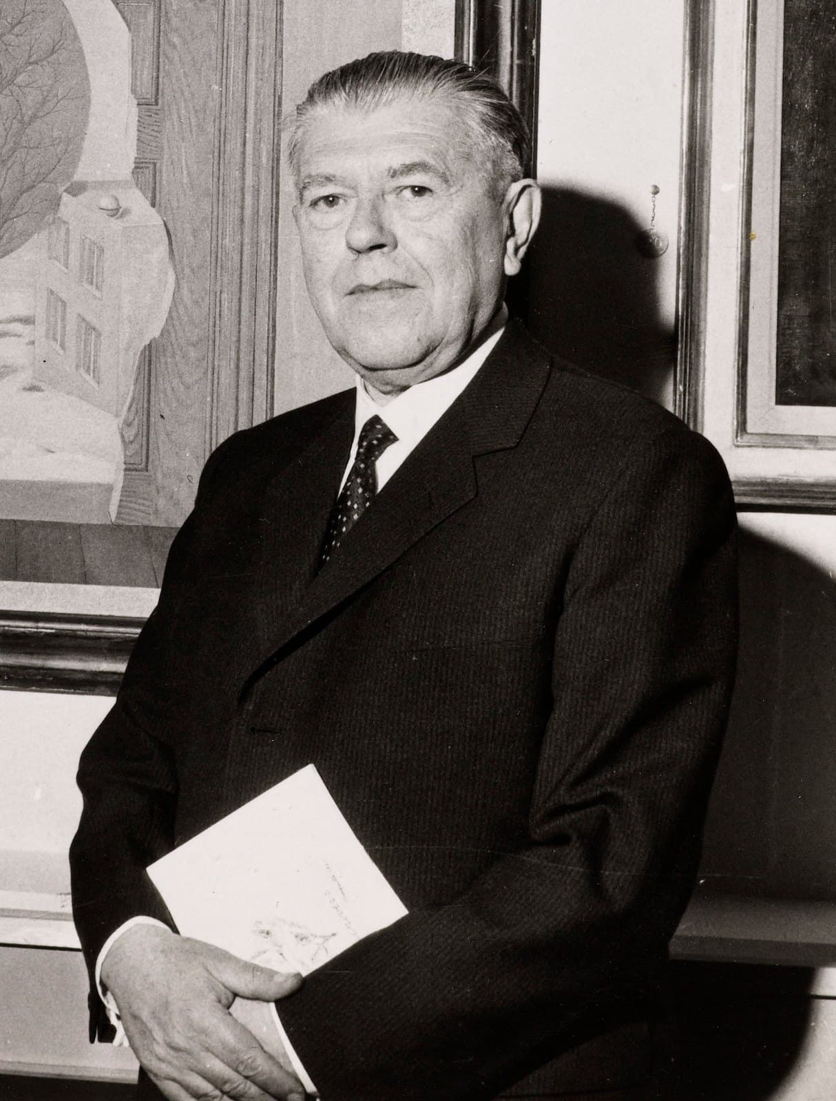 René Magritte in a dark suit standing before one of his framed paintings, holding a catalogue.