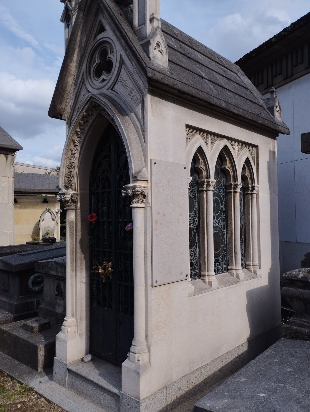 A small pale stone funerary chapel in Gothic style at Passy cemetery, with a pointed arched doorway of dark ironwork, three lancet windows of leaded glass, and a marble plaque on the side wall; the gable carries the name Pauline M. Tarn, and roses have been pushed through the ironwork.