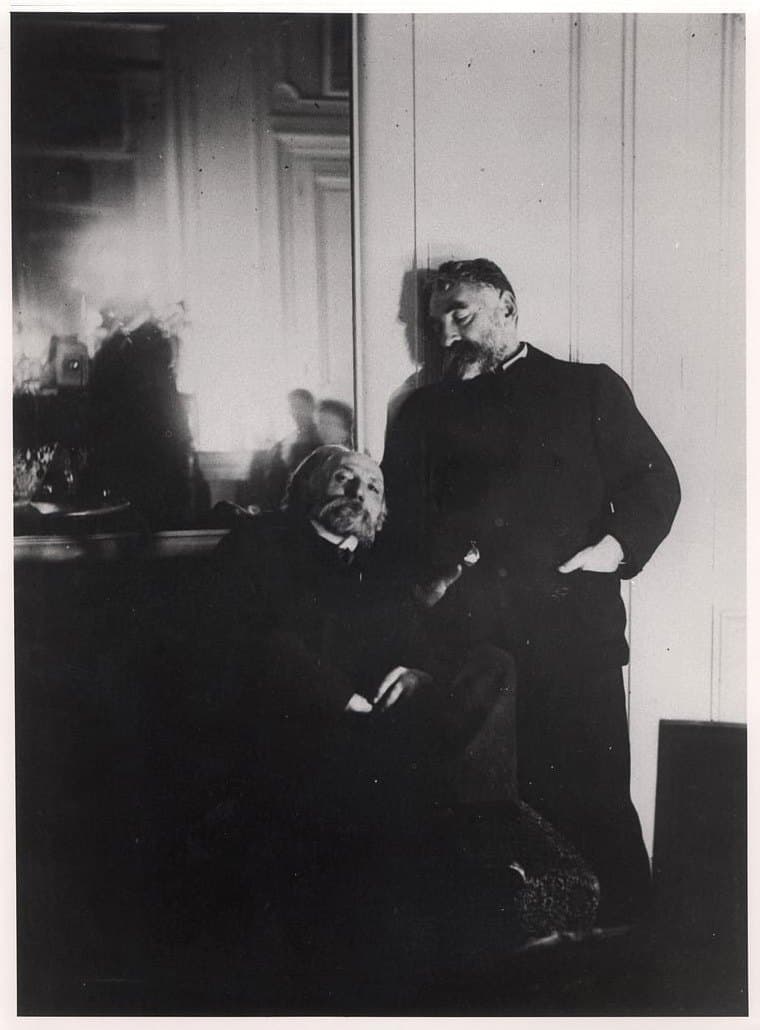 Renoir seated on a sofa beside Stéphane Mallarmé standing against a mirror, photographed by lamplight