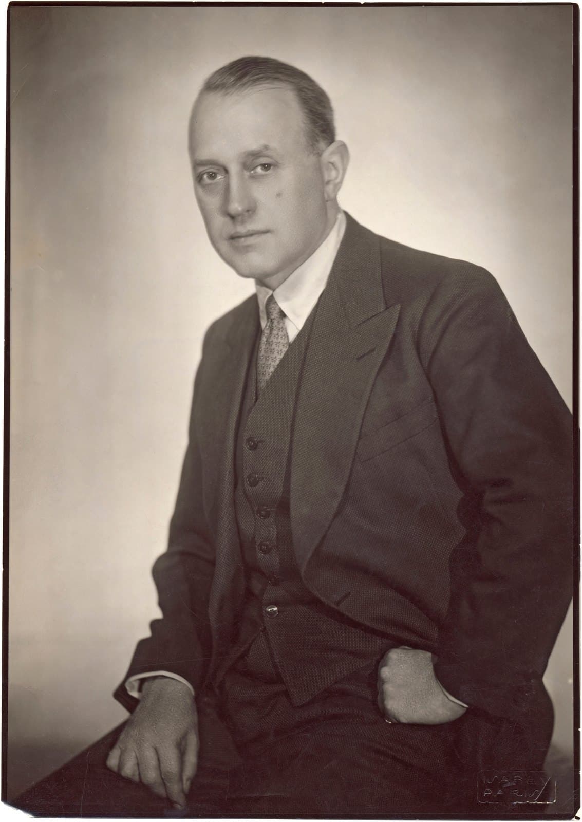 Rolf de Maré seated in a three-piece suit, one hand in his trouser pocket, in a studio portrait.