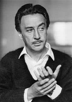 Romain Gary in a dark cardigan over an open collar, hands clasped, looking aside.