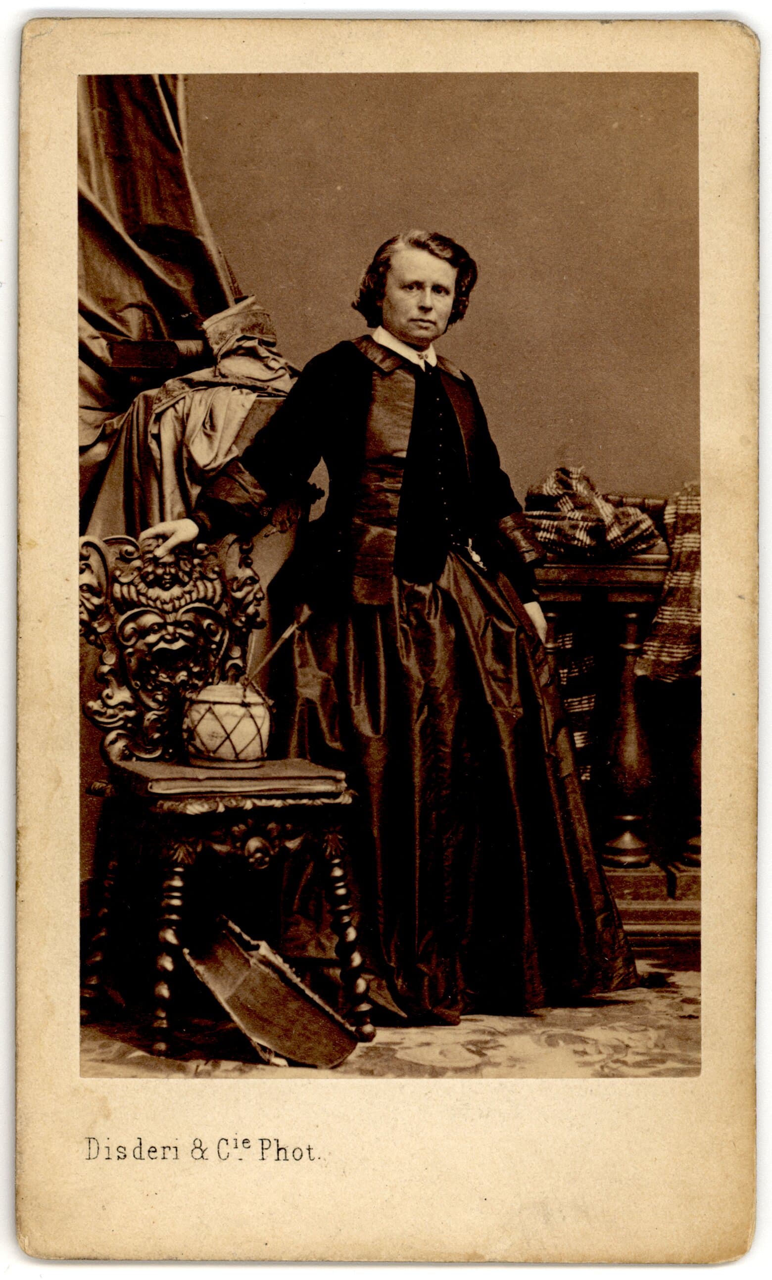 Rosa Bonheur standing in a dark dress and jacket, with one hand on an ornate chair.