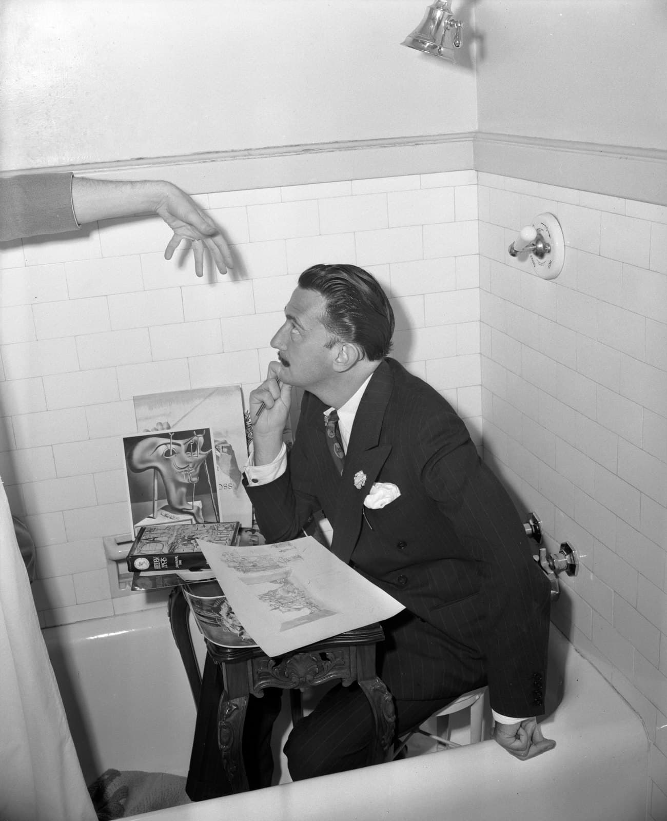 Salvador Dalí in a pinstriped suit, seated on a carved side table inside an empty bathtub, holding a drawing and looking up at a disembodied arm reaching in from the left