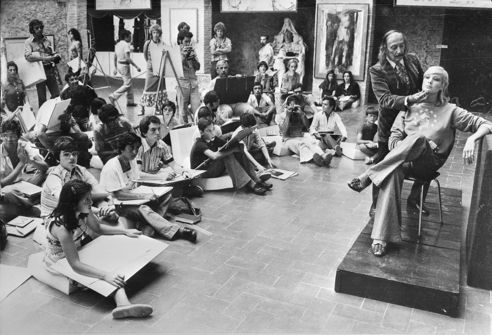 Salvador Dalí standing on a low dais beside a seated model, facing a hall of young art students sitting on the floor with drawing boards, photographers among them