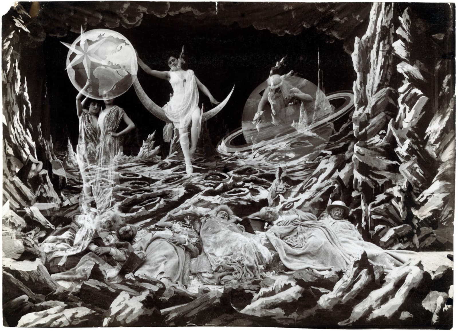 Still from A Trip to the Moon: the astronomers asleep under blankets on the crater floor while Phoebe on her crescent, a star goddess, and Saturn in his ringed planet look on