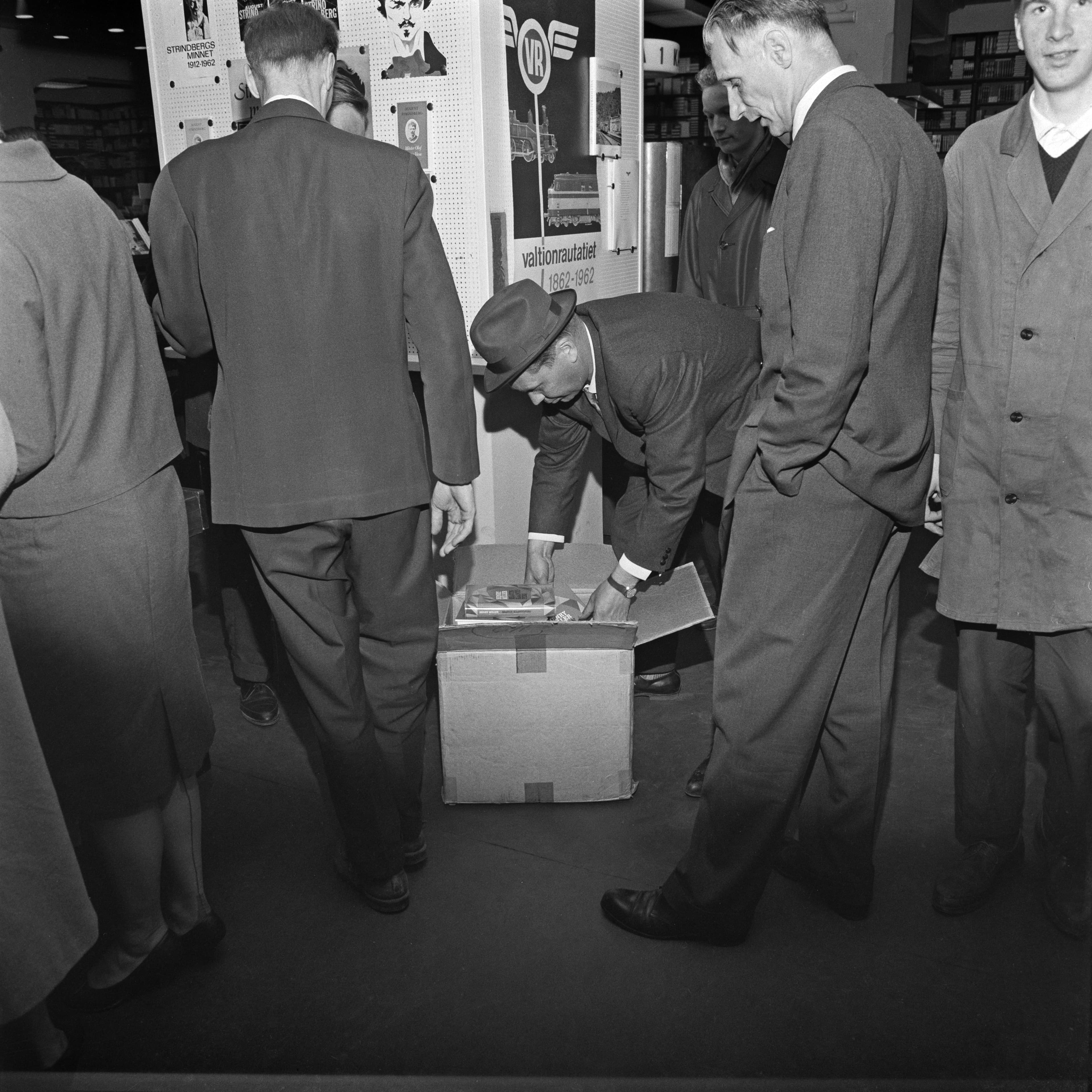Men in suits around an open cardboard box of books on a bookshop floor as one reaches in to remove copies.