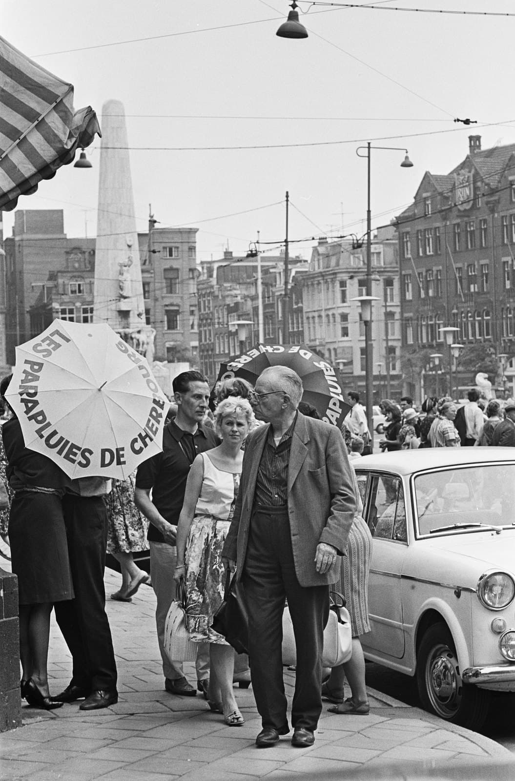 Passers-by walk past a couple kissing behind a white umbrella printed Les Parapluies de Cherbourg on a crowded Amsterdam pavement, a second dark branded umbrella behind them and the National Monument in the distance