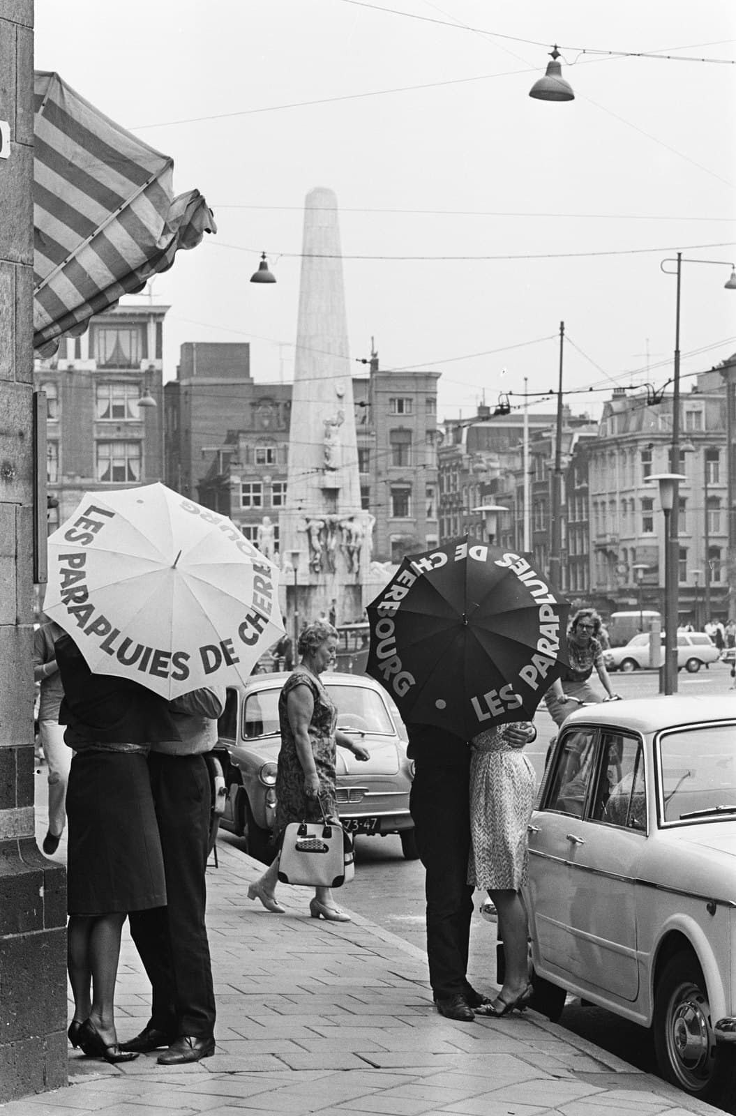 Two couples kiss on an Amsterdam pavement, each hidden behind an umbrella printed Les Parapluies de Cherbourg — one white, one black — with the National Monument on Dam Square and parked 1960s cars behind them