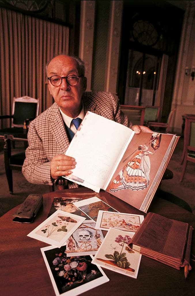 Black-and-white photograph of Vladimir Nabokov at seventy, seated outdoors in Montreux in a cardigan, hands folded, looking directly at the camera