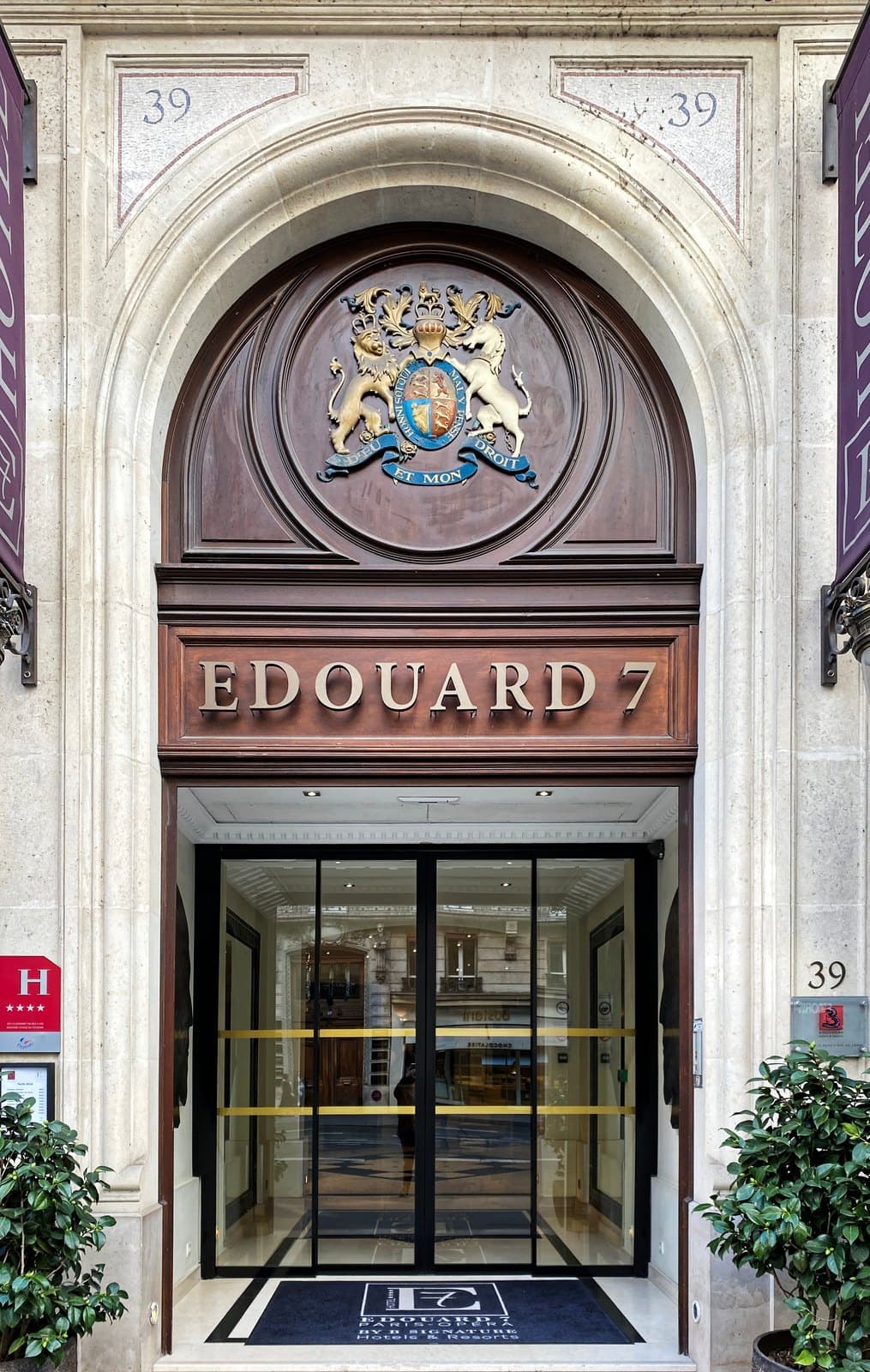 Photograph of the arched stone entrance of Hôtel Edouard 7, with the British royal coat of arms above bronze letters reading Edouard 7 and the number 39 on both sides of the arch.