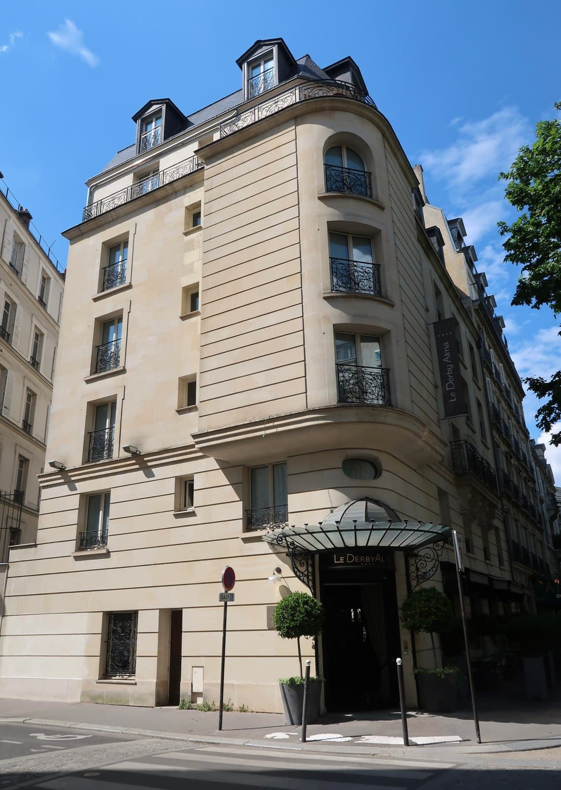 Photograph of the pale stone corner building of Le Derby Alma with a rounded corner bay, wrought-iron balconies and mansard windows, a glass-and-iron canopy lettered Le Derby Alma over the door and a vertical black banner with the hotel's name.